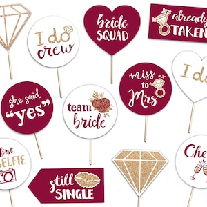May include: A collection of photo booth props for a wedding or bridal shower. The props include diamond and heart shapes, speech bubbles, and signs with phrases like "Bride Squad," "I do," and "Cheers." The props are in burgundy, white, and gold.