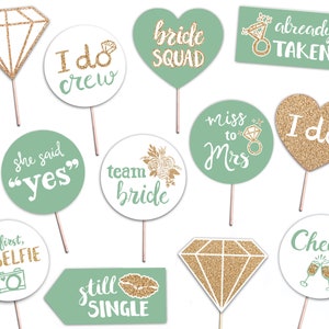 Op de afbeelding: Set van 11 groene en gouden glitter fotobooth props met zinnen zoals "bride squad", "I do crew", "she said yes", "miss to Mrs", "team bride", "but first, #selfie", "still single", "already taken", "I do", "cheers" en "diamond".