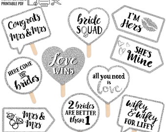 May include: Set of 10 silver glitter photo booth props with various sayings for a wedding or bridal shower. The sayings include "Congrats Mrs & Mrs", "Here Come The Brides", "Bride Squad", "Love Wins", "I'm Hers", "She's Mine", "All You Need Is Love", "2 Brides Are Better Than 1", "Mrs & Mrs", and "Wifey & Wifey For Lifey".