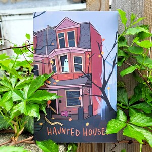 May include: A black and white illustration of a haunted house with a ghost in the window. The house is red with white trim and has a porch with a door. The text "Haunted House" is printed below the house.