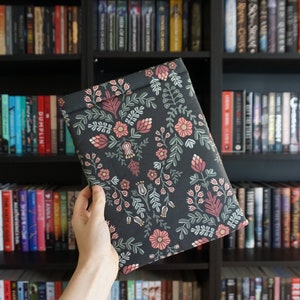 May include: A dark grey book sleeve featuring a floral pattern in pink, red, and green. The sleeve is held in front of a bookshelf filled with books. The floral design includes stylized flowers and leaves.