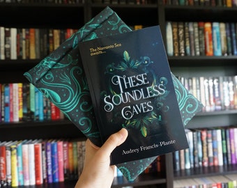 May include: A book titled "These Soundless Caves" with a dark cover and ornate lettering, held with a teal patterned book sleeve. The book and sleeve are set against a backdrop of a bookshelf filled with books.