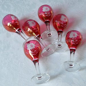 May include: Set of six red glass stemmed wine glasses with white floral etching. The glasses have a clear base and a red, iridescent body.
