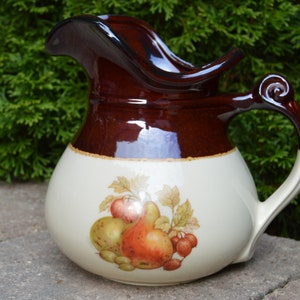 May include: A brown and white ceramic pitcher with a wide, flared spout and a handle. The pitcher features a painted design of fruit, including pears, apples, and grapes.