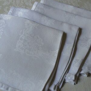 May include: A set of five white linen napkins with an embossed floral pattern. The napkins are folded and stacked on a white background.