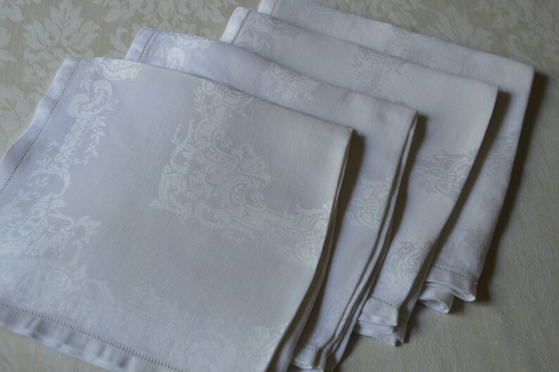 May include: A set of five white linen napkins with an embossed floral pattern. The napkins are folded and stacked on a white background.