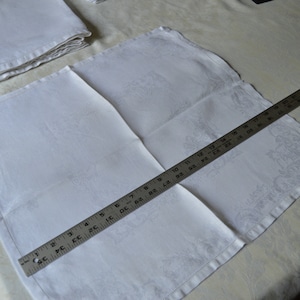 May include: A white linen napkin with a subtle embossed pattern, folded and measured with a ruler. The ruler shows 18 inches.