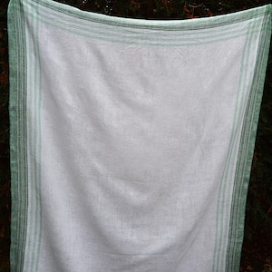 May include: A white linen tablecloth with a green border and stripes. The tablecloth is hanging outdoors, with the green border and stripes framing the white center. The fabric appears to be slightly wrinkled.
