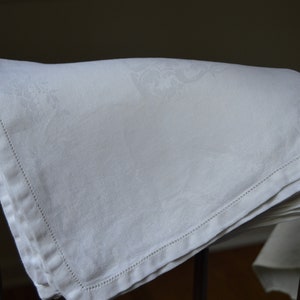 May include: A white linen tablecloth with a delicate embroidered pattern, folded and displayed on a wooden chair.