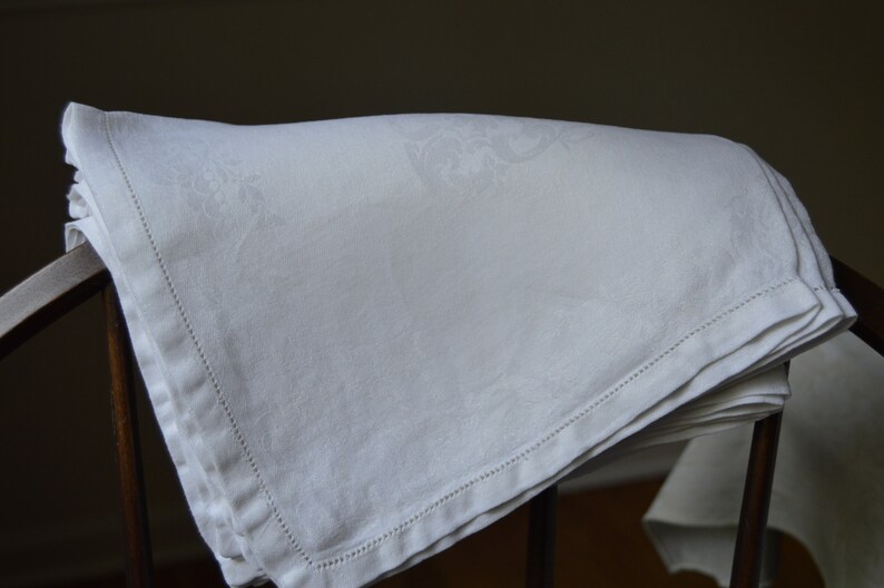 May include: A white linen tablecloth with a delicate embroidered pattern, folded and displayed on a wooden chair.