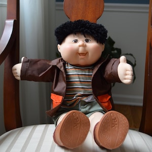 May include: A Cabbage Patch Kids doll sits on a chair. The doll has a round face, black curly hair, and is wearing a brown jacket with orange accents, a striped shirt, and green shorts. The doll's shoes are brown.