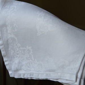 May include: White linen napkin with a delicate floral pattern. The napkin is folded and draped over a wooden chair.