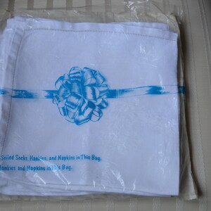 May include: A package of white linen napkins with a blue ribbon and bow. The package has the text "turn, Soiled Socks, Hankies, and Napkins in This Bag. lore Hankies and Napkins in This Bag."