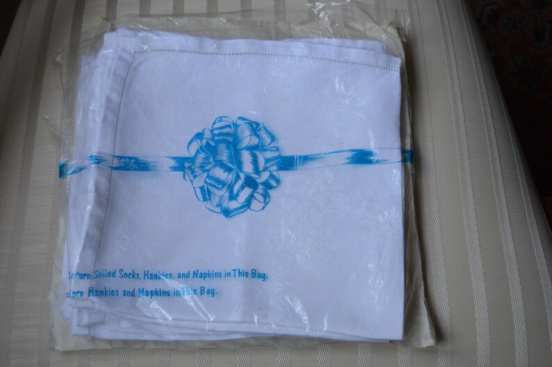 May include: A package of white linen napkins with a blue ribbon and bow. The package has the text "turn, Soiled Socks, Hankies, and Napkins in This Bag. lore Hankies and Napkins in This Bag."