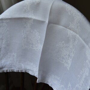 May include: White linen tablecloth with a delicate, embossed floral pattern. The tablecloth is folded in half and draped over a wooden chair.