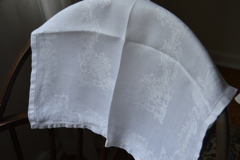 May include: White linen tablecloth with a delicate, embossed floral pattern. The tablecloth is folded in half and draped over a wooden chair.
