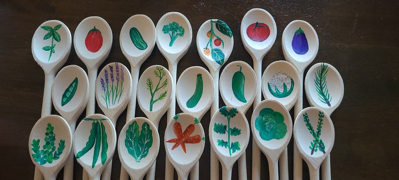May include: A collection of wooden spoons painted with various vegetables and herbs. The spoons are arranged in a row on a wooden surface. The vegetables and herbs include broccoli, carrots, peas, lavender, dill, cucumbers, tomatoes, eggplant, and more.