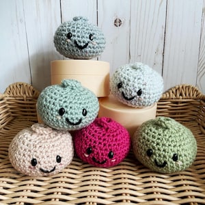 May include: A collection of six round, crocheted plush toys in various colors including light blue, sage green, beige, magenta, and speckled white. Each plush features a small top knot, black embroidered eyes, and a stitched smiling mouth.