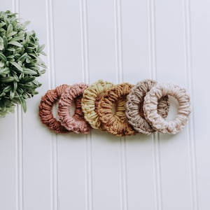 May include: A collection of six handmade scrunchies in various warm, earthy tones. The scrunchies are made of a textured yarn, with colors ranging from a deep rust to a light beige. They are arranged in a row against a white background.