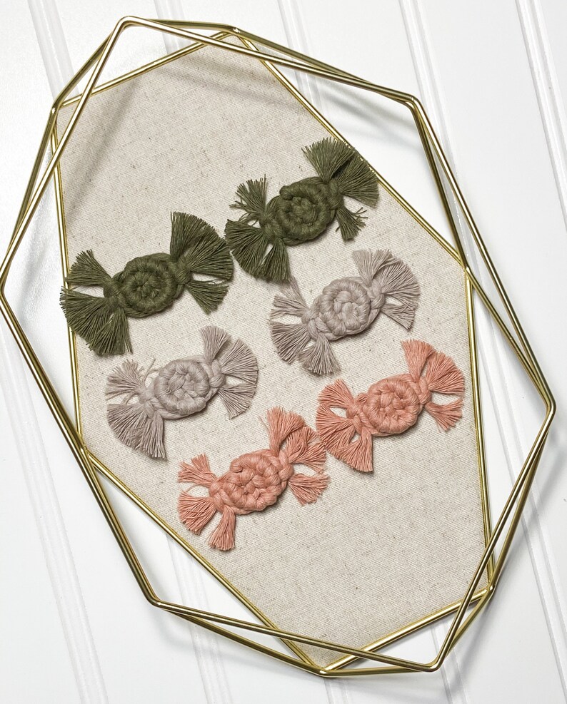 K&ouml;nnte beinhalten: Sechs handgefertigte Makramee-Ornamente in verschiedenen Farben, darunter Gr&uuml;n, Beige und Rosa. Die Ornamente werden auf einem goldenen Tablett aus Metall pr&auml;sentiert.