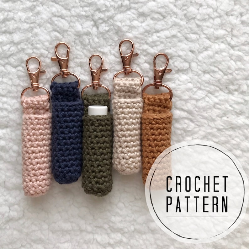 Pu&ograve; includere: Cinque porta-balsamo per labbra all'uncinetto con portachiavi rosa, blu navy, verde oliva, crema e marrone. I porta-balsamo sono appesi su una superficie bianca e soffice. Il testo "CROCHET PATTERN" &egrave; scritto in un cerchio sotto i porta-balsamo.