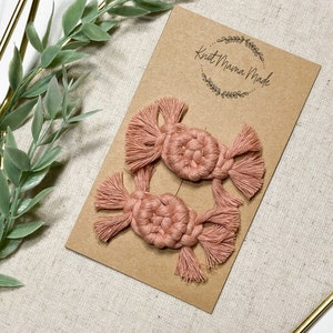 K&ouml;nnte beinhalten: Zwei rosa Makramee-Haarklammern mit Quasten. Die Klammern werden auf einer braunen Karte mit dem Text "Knot Mama Made" in einem Kreis angezeigt.