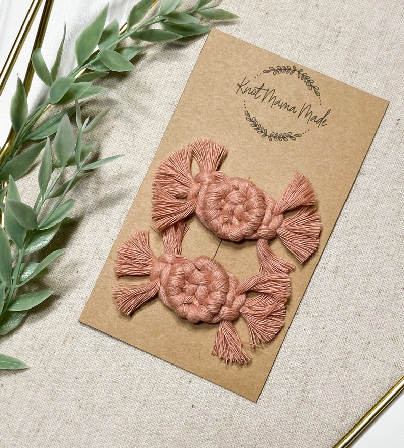 K&ouml;nnte beinhalten: Zwei rosa Makramee-Haarklammern mit Quasten. Die Klammern werden auf einer braunen Karte mit dem Text "Knot Mama Made" in einem Kreis angezeigt.