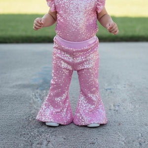 May include: A pink sequined outfit for a baby or toddler, featuring a top with ruffled sleeves and flared pants. The outfit is covered in shimmering sequins, creating a sparkly effect. The pants have a wide-leg design. The outfit is worn with white shoes.