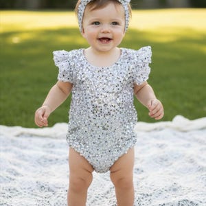 May include: A silver sequined romper featuring short ruffled sleeves and a round neckline, paired with a matching silver sequined headband adorned with a large bow. The garment is covered in shimmering metallic sequins that reflect light.
