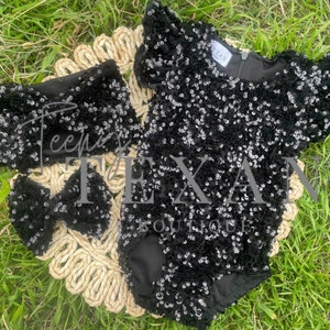 May include: A black sequin bodysuit with ruffle sleeves and a matching headband and bow. The headband and bow are also covered in black sequins.