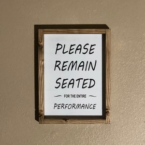 May include: A wooden framed sign with the text "PLEASE REMAIN SEATED FOR THE ENTIRE PERFORMANCE" in black lettering on a white background.