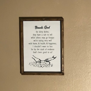 May include: A framed sign with a white background and a dark brown wooden frame. The sign features the text "Thank God for dirty dishes; they have a tale to tell..." in black font. A graphic of hands washing dishes is at the bottom.