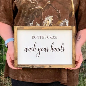 May include: A rectangular wooden sign with a white background and black text that reads "DON'T BE GROSS wash your hands." The sign has a rustic, light brown frame. The sign is being held by a person.