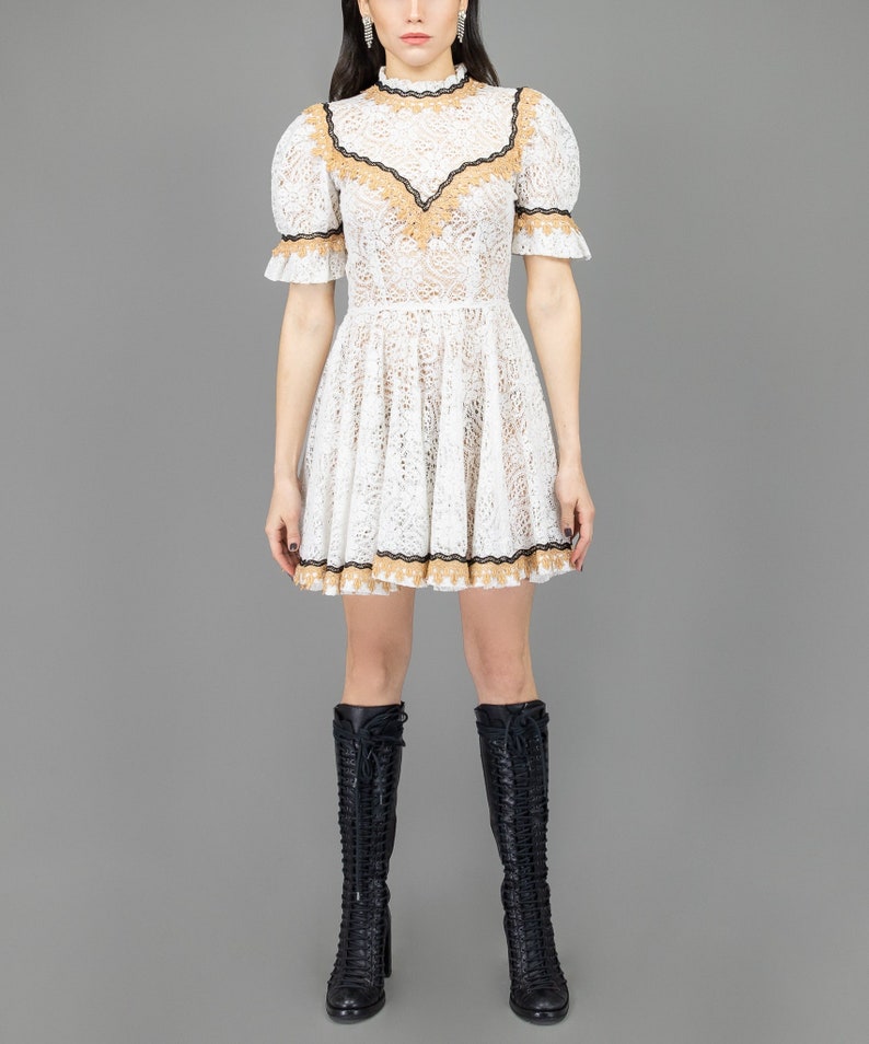 May include: A white lace dress with a high neckline and short sleeves. The dress has a brown and black trim around the neckline and hem. The dress is fitted at the waist and flares out to a mini length.