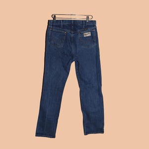 May include: A pair of dark blue denim jeans with a classic straight leg fit. The jeans have a leather patch on the back waistband with the Wrangler logo.