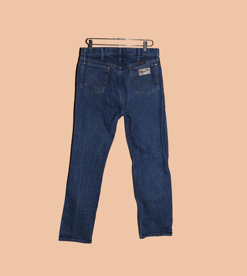 May include: A pair of dark blue denim jeans with a classic straight leg fit. The jeans have a leather patch on the back waistband with the Wrangler logo.