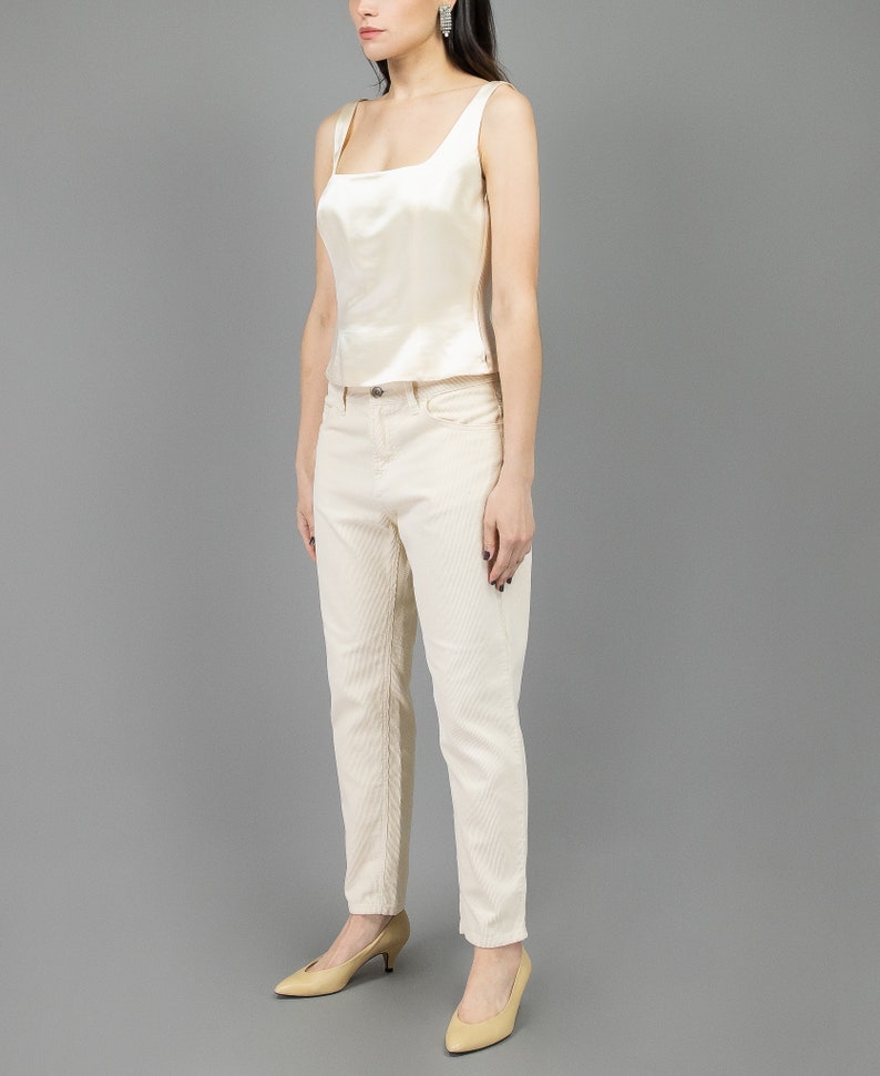 collector of vision model is wearing a size medium cream escada silk corset showing off the side with square neckline, thick straps, styled with cream corduroy pants, nude low pumps, and crystal dangling earrings