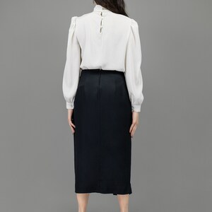 collector of vision model is wearing a size medium black short long midi skirt showing off the back with back zipper closure, long midi length, styled with a formal white shirt, plum buckle pumps, and crystal dangling earrings