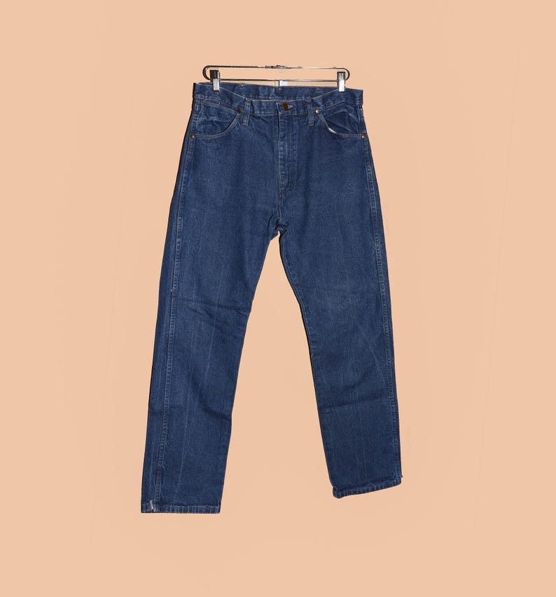 May include: A pair of dark blue denim jeans hanging on a hanger. The jeans have a classic straight leg style and a button fly.