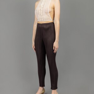 May include: A white lace halter top with a high neckline and a tie at the waist. The top is worn with brown pleated trousers and beige heels.