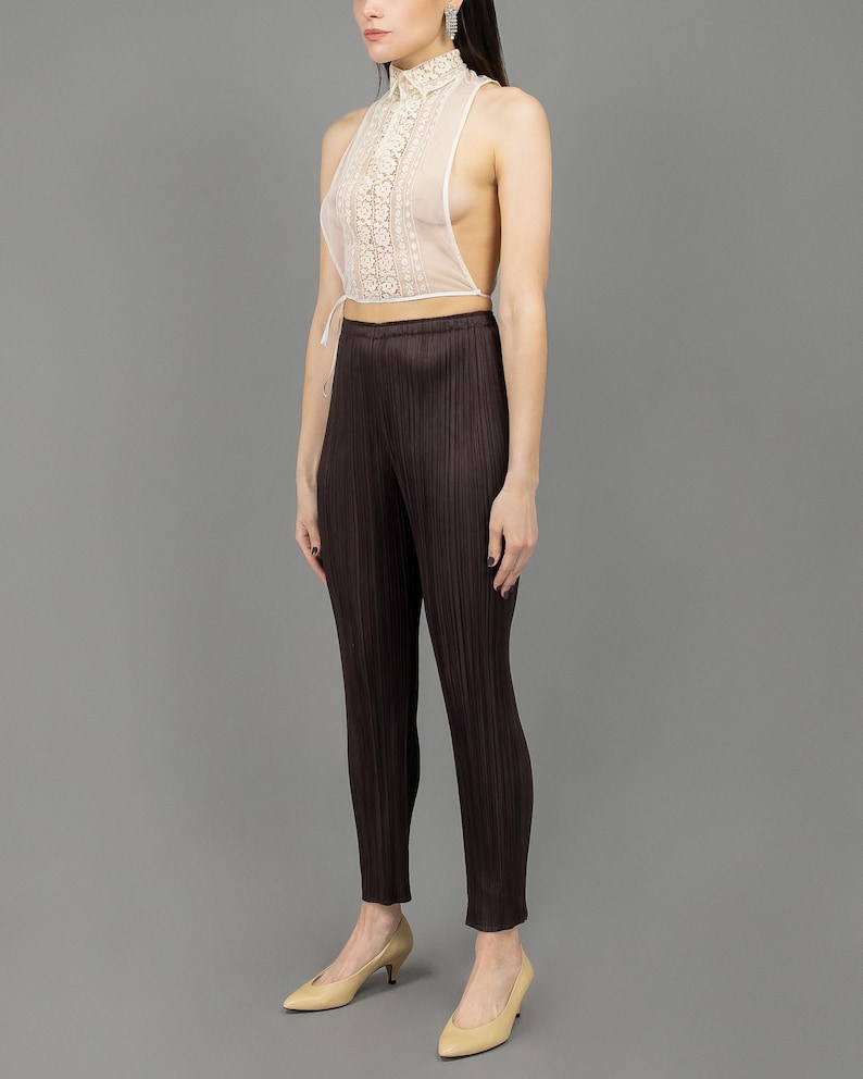 May include: A white lace halter top with a high neckline and a tie at the waist. The top is worn with brown pleated trousers and beige heels.