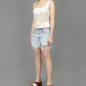 collector of vision model is wearing a size medium cream escada silk corset with scoop neckline, thick straps, styled with cut off levis blue jean shorts, 70s red and white clogs, and crystal dangling earrings