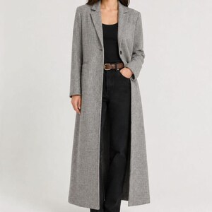 May include: A long, grey, textured wool-blend coat with notched lapels and a single-button closure. Underneath, a black sleeveless top is tucked into black trousers secured with a brown leather belt featuring a gold-toned rectangular buckle.