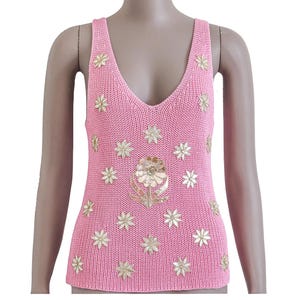 K&ouml;nnte beinhalten: Ein rosa gestricktes Tanktop mit einem gro&szlig;en goldenen Blumendesign in der Mitte und kleineren goldenen Blumenakzenten auf der gesamten Oberfl&auml;che.