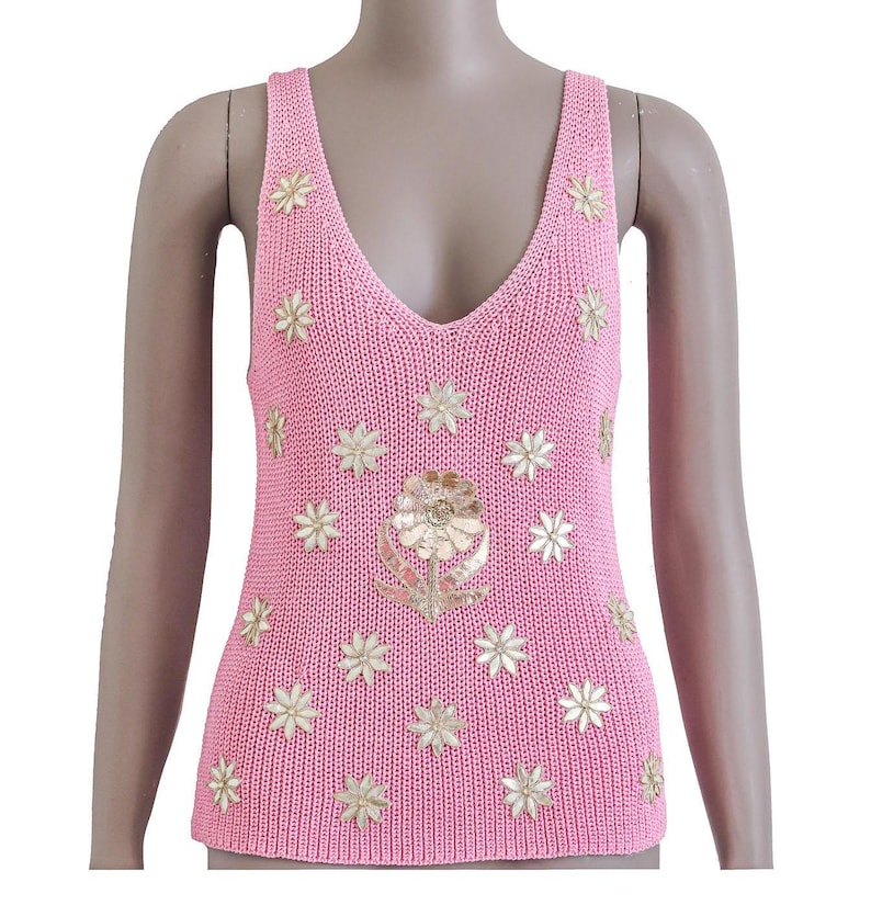 K&ouml;nnte beinhalten: Ein rosa gestricktes Tanktop mit einem gro&szlig;en goldenen Blumendesign in der Mitte und kleineren goldenen Blumenakzenten auf der gesamten Oberfl&auml;che.