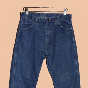 May include: A pair of dark blue denim jeans hanging on a silver hanger. The jeans have a classic five-pocket style and a straight leg.