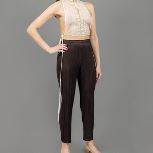 May include: A white lace halter top with a high neck and a tie at the back. The top is worn with brown pleated trousers and beige heels.