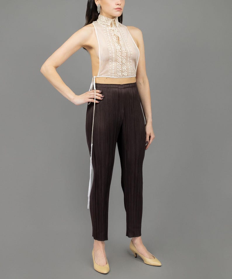 May include: A white lace halter top with a high neck and a tie at the back. The top is worn with brown pleated trousers and beige heels.