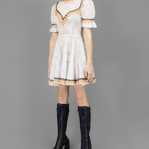collector of vision model is wearing a size small white lace baby doll dress showing the side with western gold and black trim, mock neckline, short puff sleeves, fit and flare ruffle hemline, styled with black tall boots and crystal dangling earring
