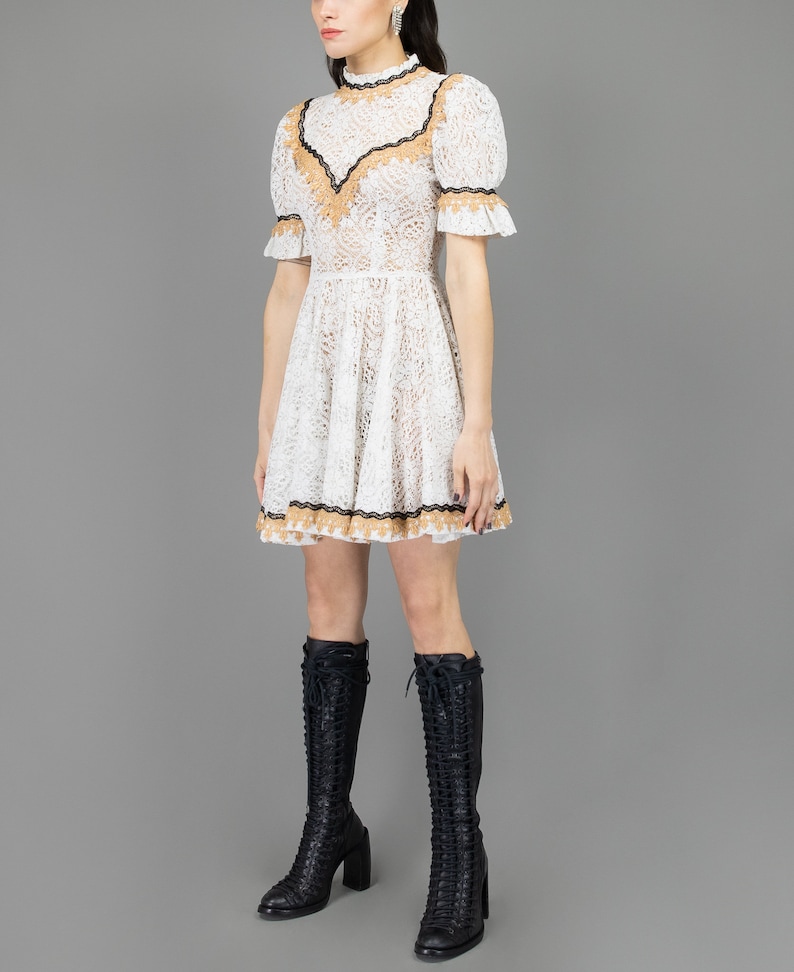 collector of vision model is wearing a size small white lace baby doll dress showing the side with western gold and black trim, mock neckline, short puff sleeves, fit and flare ruffle hemline, styled with black tall boots and crystal dangling earring